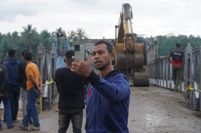 
					Seorang warga memberi kabar kepada kerabatnya lewat panggilan video tentang akses jembatan Bailey yang menghubungkan Bireuen dengan Kabupaten Bener Meriah sudah dapat dilalui di atas Sungai Teupin Mane, Kabupaten Bireuen, Aceh, Selasa (16/12/2025). Jembatan sepanjang sekitar 180 meter ini kembali menghubungkan akses darat Bireuen–Aceh Tengah yang terputus akibat banjir bandang, sekaligus memperlancar mobilitas warga dan distribusi logistik pascabencana. Infopublik